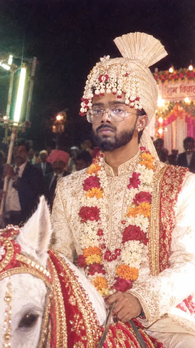 A groom in a cream sherwani and turban on a decorated white horse at a 1986 night wedding procession, lit by hard direct flash