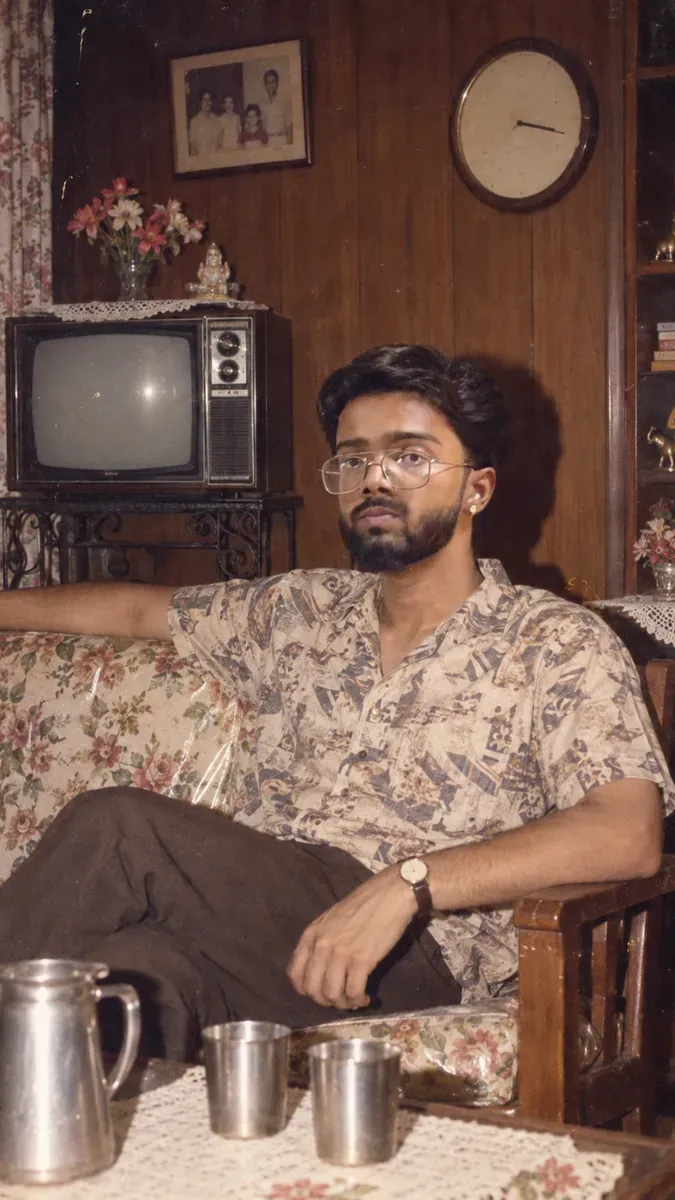 A person seated on a floral sofa in a wood-panelled 1986 Indian drawing room beside a boxy television, lit by a direct camera flash