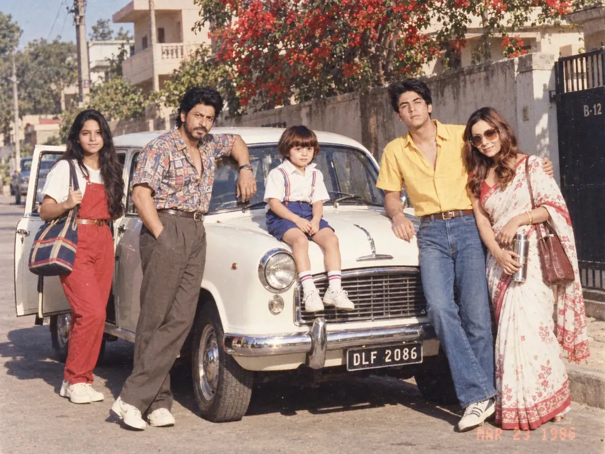 A family posed around a white Ambassador on an Indian street in bright 1986 morning sun, dressed in Sunday best