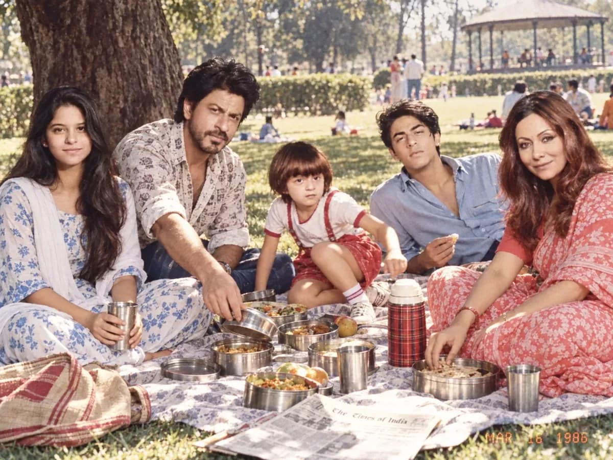 A family sitting on a cotton sheet under a tree in an Indian park in 1986 with steel tiffin boxes, in dappled sunlight